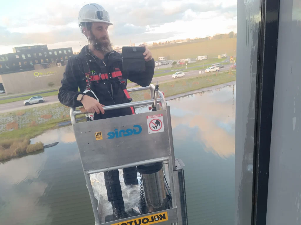 Poseur Easy Film en harnais dans une nacelle à mât vertical, appliquant un film anti UV sur un vitrage à 11 mètres au-dessus d’un plan d’eau pour SNEF